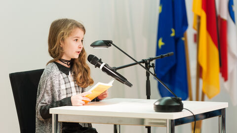 Vorlesewettbewerb 2019 Ein Mädchen mit blonden Haaren und einem grauen Oberteil sitzt an einem Tisch mit einem Buch in der Hand. Sie spricht in ein Mikrofon.