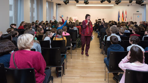 Vorlesewettbewerb 2019 Eine Frau läuft durch einen Gang zwischen Stuhlreihen mit einem Mikrofon in der Hand. Auf den Stühlen sitzen Kinder, die teilweise die Hand haben.