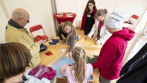 Acht Personen stehen gemeinsam um einen Tisch und verschieben geometrische Figuren: Mathematik zum Anfassen. Der Knobel- und Spielstand des Gießener Museums „Mathematikum“
