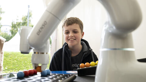 Ein kleiner Junge beobachtet einen Roboterarm beim erfolgreichen Einsetzen von bunten Kugeln in eine Lochplatte: …Boah, der hat es echt geschafft…?!...