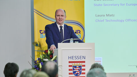 Europastaatsekretär Uwe Becker begrüßt die Gäste Europastaatsekretär Uwe Becker begrüßt die Gäste: Er steht am Rednerpult und spricht zum Publikum