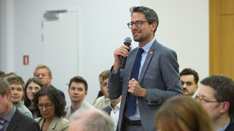 Fragen aus dem Publikum - im Foto zu sehen ist ein Fragesteller im Anzug, der freundlich lächelnd ins Mikrofon spricht und zur Bühne schaut