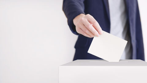 Hand mit Stimmzettel, der in eine Wahlurne geworfen wird.