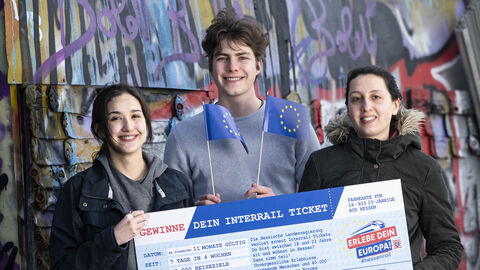 Drei junge Menschen stehen vor einer Graffiti besprühten Wand mit einem "HessenRail"-Scheck in Händen. Die mittlere Person wedelt mit kleinen Europa-Flaggen