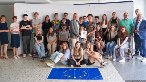 Europaministerin überreicht Zuwendung in Höhe von 1.150 Euro an Förderverein der Wölfersheimer Schulen e.V. Europaministerin Puttrich mit mehreren Beteiligten Personen und Schülerinnen und Schülern in einem Raum der Schule. Sie stehen in drei Reihen hintereinander, Frau Puttrich hält den Bescheid in Händen.