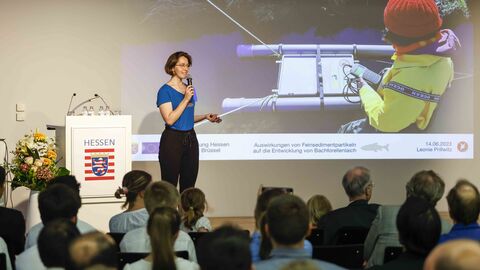 Eine Frau steht mit Mikrofon auf der Bühne, erläutert ein Projekt zu Bachforellenlaich