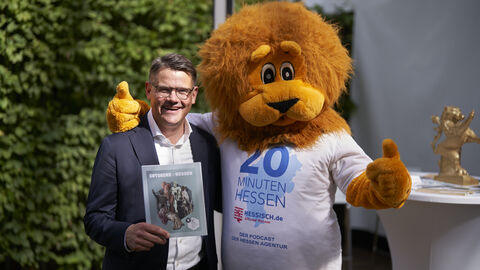 Ministerpräsident Boris Rhein steht links neben einem lebensgroßen Hessen-Löwen-Maskottchen im Freien im Sonnenschein