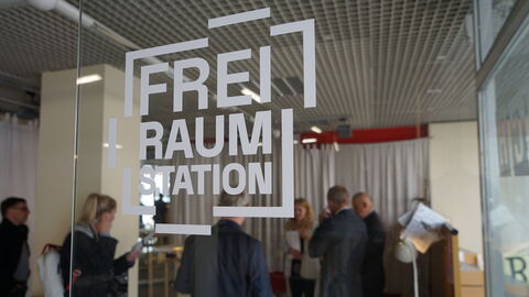 Der Marktplatz der Kleinstadt ist der Ausgangspunkt des Projektes, um ihn herum gibt es nun den Co-Working-Space FachWerkerei, einen gemeinschaftlichen Garten, die Gemeinschaftsküche und das MachWerk, eine offene Quartierswerkstatt. Blick durch eine Glasscheibe mit der Aufschrift „FREIRAUMSTATION“ auf den Chef der Hessischen Staatskanzlei, Staatsminister Axel Wintermeyer, sowie seine Gesprächspartnerinnen und -partner