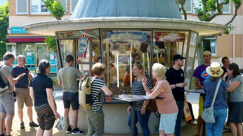 Mehrere Personen stehen vor einem kleinen Kioskhäuschen an Stehtischen.