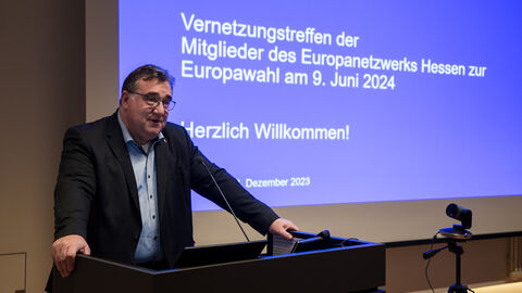 Regierungspräsident Mark Weinmeister begrüßt die Mitglieder des Multiplikatorentreffens. Regierungspräsident Mark Weinmeister steht vor einer Leinwand, auf der die erste Folie einer Präsentation zu sehen ist.