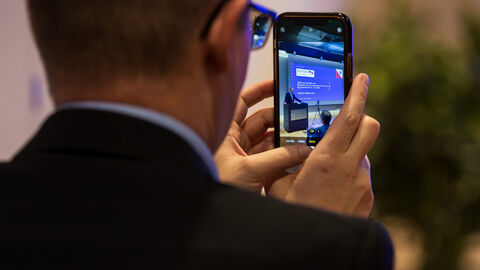 Handyaufnahme von Europastaatssekretär Uwe Becker am Rednerpult Es ist ein Mann von hinten zu sehen, der mit seinem Smartphone den Redner auf der Bühne fotografiert.