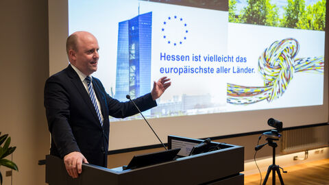 Europastaatssekretär Uwe Becker erläutert die Aktivitäten der Hessischen Landesregierung anlässlich der Europawahl Europastaatssekretär Uwe Becker steht am Rednerpult mit erhobener Hand; im Hintergrund ist die Präsentation zu sehen.