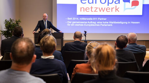 Europastaatssekretär Uwe Becker erläutert die Aktivitäten der Hessischen Landesregierung anlässlich der Europawahl Es sind Europastaatssekretär Uwe Becker von vorne und das Publikum von hinten zu sehen. Auf der Leinwand im Hintergrund informiert eine Folie über das Europanetzwerk.