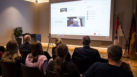 Auf einer Leinwand ist Oliver Hänsgen zu sehen, im Vordergrund sieht man ein Teil des Publikums von hinten. 