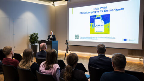 Patrycja Sypel, Referentin für Kommunikation und Öffentlichkeitsarbeit in der Vertretung der Europäischen Kommission in Deutschland, erläutert die Aktivitäten der Kommission anlässlich der Europawahl Patrycja Sypel von der Europäischen Kommission ist am Rednerpult, im Hintergrund ist ihre Präsentation und im Vordergrund das Publikum von hinten zu sehen.