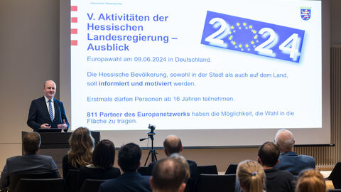 Europastaatssekretär Uwe Becker erläutert die Aktivitäten der Hessischen Landesregierung anlässlich der Europawahl Es sind Europastaatssekretär Uwe Becker, im Hintergrund eine Präsentation auf einer Leinwand und im unteren Bildausschnitt das Publikum von hinten zu sehen.
