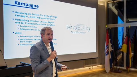 Michael Aufenanger, Europe Direct NordOstHessen, erläutert die geplanten Aktivitäten seiner Institution Michael Aufenanger ist mit dem Mikrofon abgebildet, im Hintergrund ist eine Präsentation auf einer Leinwand zu sehen.