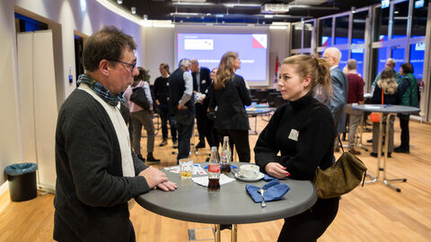 Austausch der Mitglieder des Europanetzwerks Ein Mann und eine Frau stehen an einem Stehtisch im Veranstaltungsraum und unterhalten sich, im Hintergrund sind weitere Menschen zu sehen.