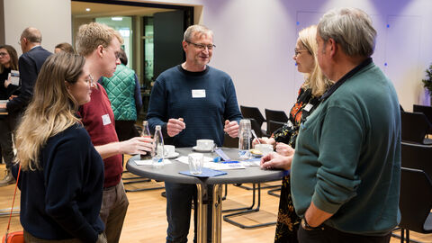 An einem Stehtisch stehen drei Männer und zwei Frauen und unterhalten sich.