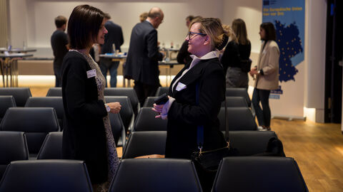 Zwei Frauen, die im Profil zu sehen sind, stehen in einer Stuhlreihe des Publikums und unterhalten sich.