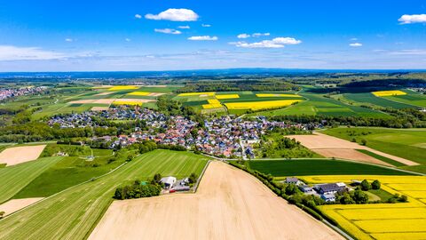 Schrägluftbild einer Kommune im hessischen Hochtaunuskreis. Neben grünen Feldern sind gelbe Rapsfelder und der blaue Himmel zu sehen.