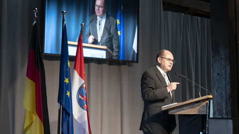 Ein Mann steh auf einer Bühne am Rednerpult. Im Hintergrund sind Flaggen aufgestellt und ein monitor überträgt seine Rede.