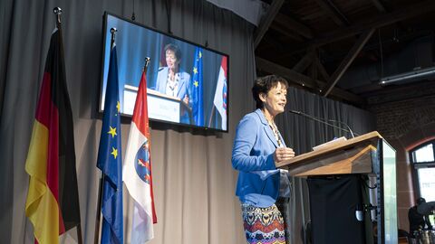 Europaempfang Europaministerin Puttrich steht auf einer Bühne am Rednerpult. Hinter ihr sind Flaggen aufgestellt und ein Monitor überträgt ihre Rede.