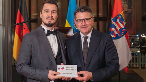 Auszeichnung für Evgenij Voznyuk Ministerpräsident Boris Rhein (rechts) neben Evgenij Voznyuk. Sie halten gemeinsam die Auszeichnung "Menschen des Respekts"  in den Händen. Im Hintergrund ein Rednerpult und die Flaggen von Deutschland, Ukraine und Hessen.