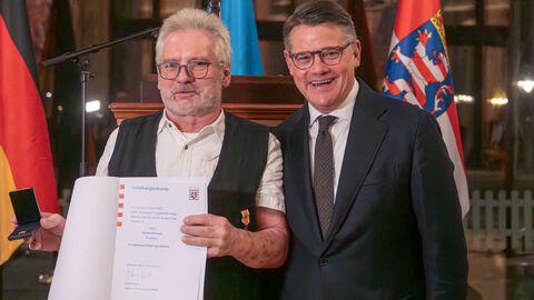 Ministerpräsident Boris Rhein und Gerhard Kimpel schauen in Kamera, im Hintergrund befinden sich 3 Flaggen. 
