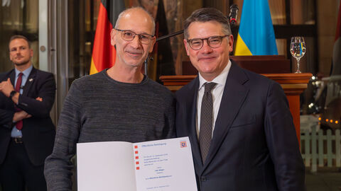 Ministerpräsident Boris Rhein und Udo Stäger aus Herborn schauen in Kamera. Im Hintergrund sieht man Flaggen. 