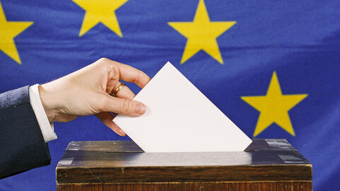 Eine Hand steckt einen Brief in eine Wahlurne, im Hintergrund ist die EU-Flagge zu sehen.