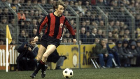 Bernd Hölzenbein auf dem Fußballfeld in einem voll besetzten Stadion, neben ihm der Fußball auf dem Rasen.