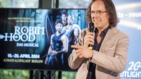 Mann spricht am Mikrofon zum Publikum: Produzent Peter Scholz berichtet von seinen Musical Projekten und hebt die gute Vernetzung von Politik und Wirtschaft vor Ort hervor.