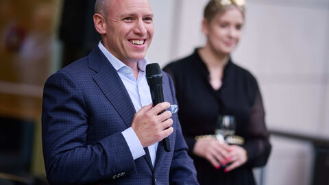 Hessens Minister für Bundesangelegenheiten, Manfred Pentz, begrüßte die Gäste aus Politik, Gesellschaft und Gastronomie zur Schlenderweinprobe 2024 Manfred Pentz hält ein Mikrofon in der Hand. Im Hintergrund steht eine weitere Person.
