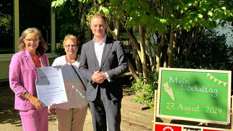 Staatskanzleichef Kuhn steht mit Schulleiterin Marika Rohrmoser und Dezernentin Anneliese Zimmer vor dem Schulgebäude. Neben ihnen eine Tafel mit er Aufschrift "Mein erster Schultag, 27. August 2024". 