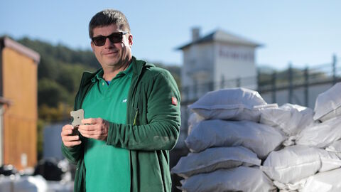 Auf dem Bild ist ein Mann mit Jeans, grünem Hemd und Sonnenbrille zu sehen, der neben einer Palette Zement steht.