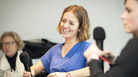 Frau im blauen Kleid lächelt in die Kamera und spricht in ein Mikrofon, welches sie in der Hand hält