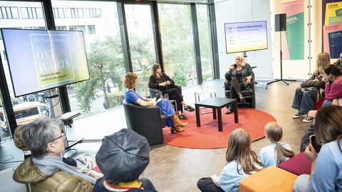 Drei Frauen im Podiums-Gespräch miteinander, im Halbkreis davor sitzen Gäste und hören zu