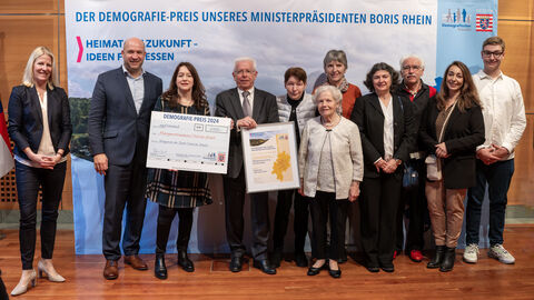 3. Preis: Magistrat der Stadt Oestrich-Winkel, "Soziale Anlauf- und Begegnungsstätte für alle Generationen – DAS INNOVATIVE SOZIALZENTRUM" Eine Gruppe Personen steht vor einem Aufsteller mit der Aufschrift "Der Demografie-Preis unseres Ministerpräsidenten Boris Rhein". Eine Person hält eine gerahmte Urkunde in Händen, eine weitere einen Scheck.