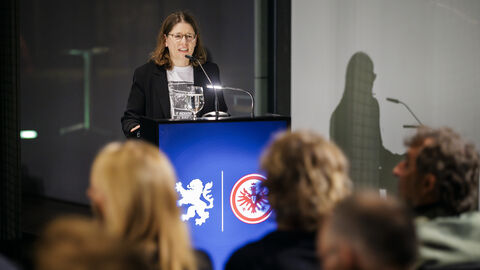 Eine Frau steht am Pult und spricht zu den Gästen der Lesung