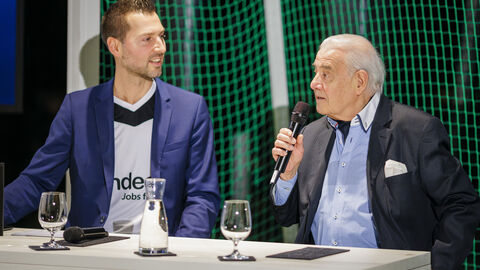 Zwei Männer sitzen nebeneinander am Tisch und sprechen miteinander 