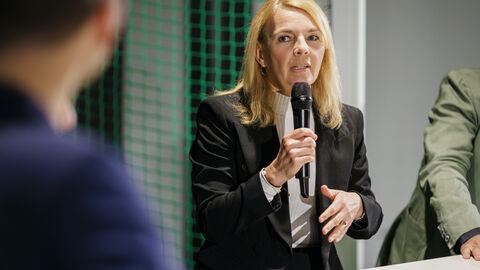 Eine Frau sitzt mit Mikrofon am Tisch und spricht mit den anderen Teilnehmern