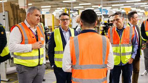 Ministerpräsident Rhein steht mit weiteren Personen in einer Lagerhalle. Sie tragen alle Warnwesten und sind ins Gespräch vertieft.