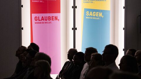 Zwei Farbige Plakate (rot-pink und gelb-blau) schmücken die Wände des Zuschauerraums.Aufschrift: Nur in einer Demokratie kannst Du Glauben, woran Du willst / Sagen, was Du willst.  