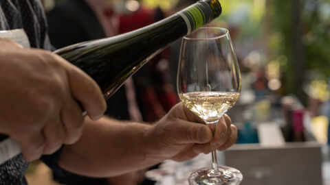 Eine Hand hält ein Weinglas, die andere Hand schenkt Weißwein ein.