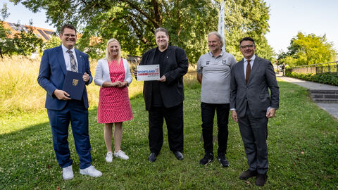 Fünf Menschen stehen nebeneinander auf einer Wiese. Die mittlere Person hält einen Zuwendungsbescheid in der Hand.