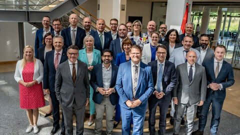 27 Männer und Frauen stehen in Anzügen und Hosenanzügen in einer Eingangshalle.