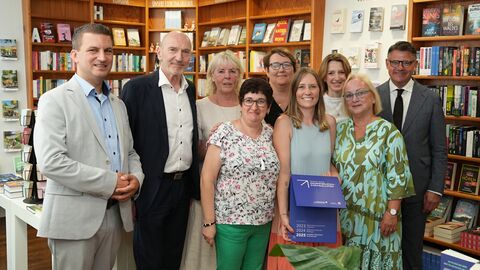 Ministerpräsident Boris Rhein und Lothar Wekel, Vorsitzender des Börsenvereins des Deutschen Buchhandels – Landesverband Hessen, Rheinland-Pfalz, Saarland e.V., überreichen Elena Rakowitz, Geschäftsführerin der Umstädter Bücherkiste, den Wanderpokal für Hessens beste Dorfbuchhandlung. Mehrere Personen stehen nebeneinander in einer Buchhandlung. Eine Person hält lächelnd eine Auszeichnung in der Hand.