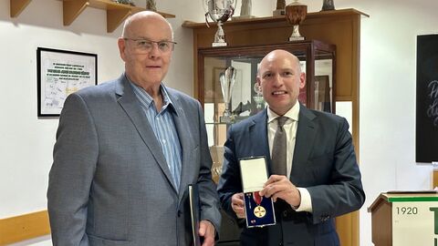 Zwei Männer stehen nebeneinander, einer hält einen Orden in den Händen.