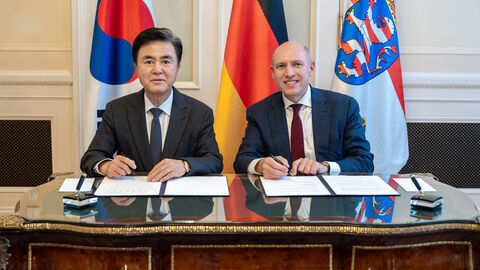 Zwei Männer sitzen am Schreibtisch. Im Hintergrund sind die koreanische, die deutsche und die hessische Flagge zu sehen.